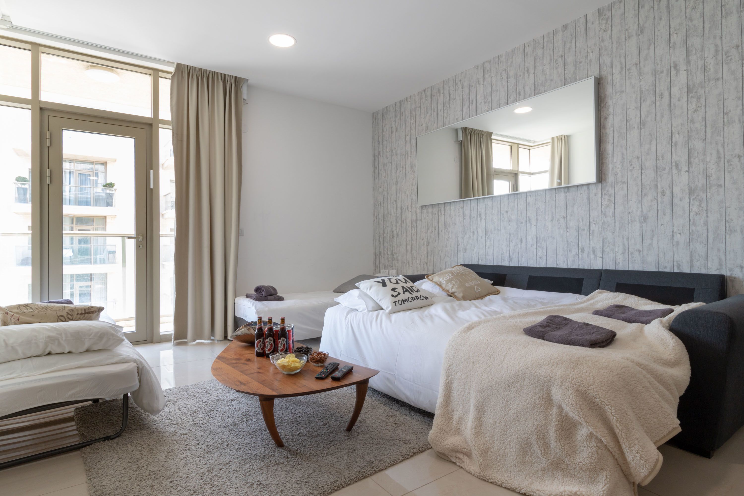 Un dormitorio moderno con una cama grande, un sofá, una mesa de centro con bebidas y aperitivos, y vistas al exterior a través de un ventanal.