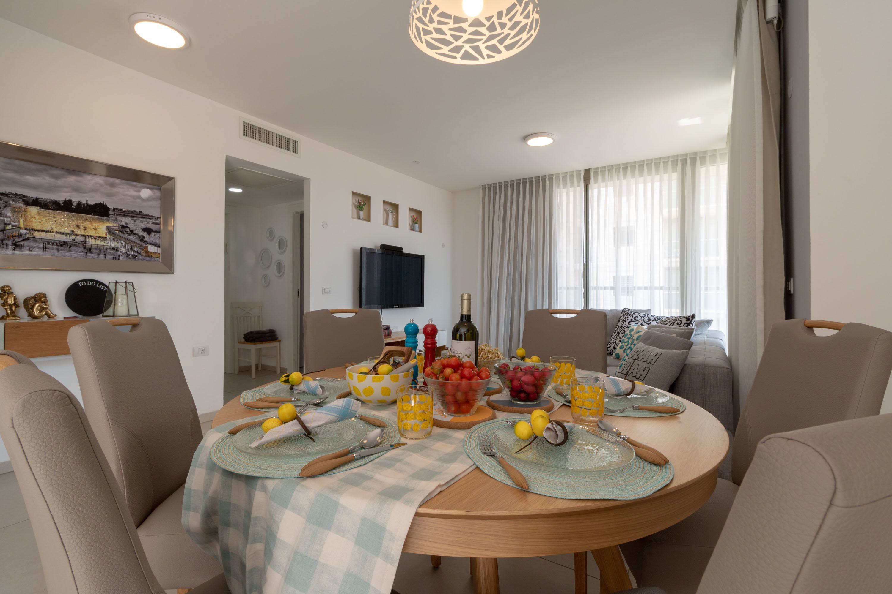 A dining area with a round table set for four, surrounded by chairs. There are bowls of fruit, glasses of juice, and a bottle of wine on the table. The room has a modern design with a large painting on the wall and a television mounted above the table.