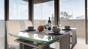 Uma mesa de jantar com tampo de vidro, rodeada por cadeiras brancas, posta com tigelas, copos e uma garrafa.