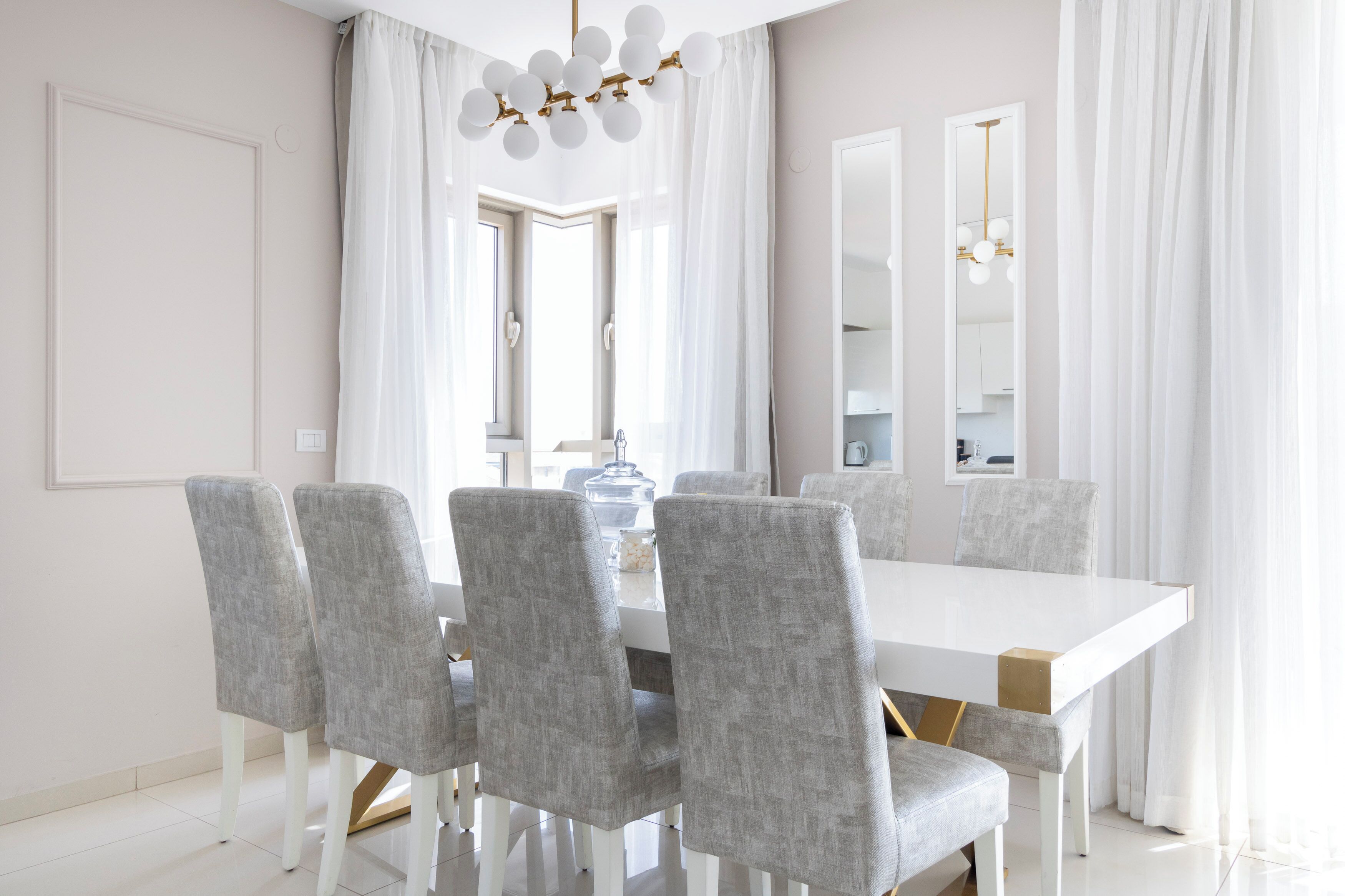 A modern dining area with a white table and grey upholstered chairs, a chandelier, and large windows with sheer curtains.