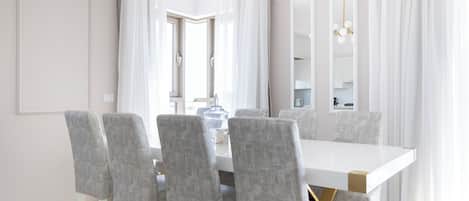 A modern dining area with a white table and grey upholstered chairs, a chandelier, and large windows with sheer curtains.