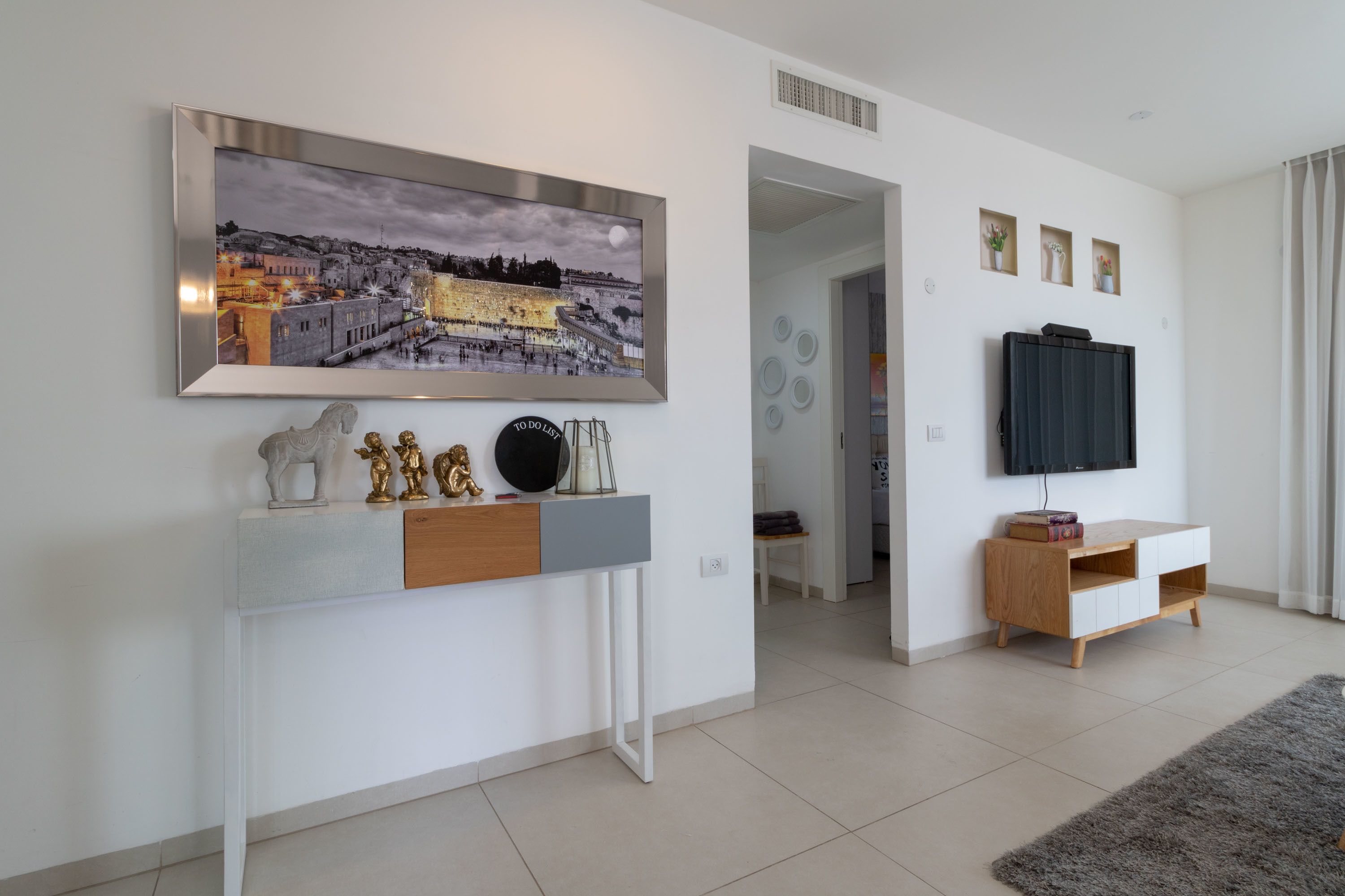 Una sala de estar moderna con un cuadro grande de un paisaje urbano enmarcado, un televisor montado y una consola decorada con objetos ornamentales.