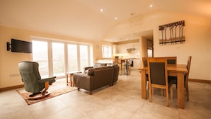 A spacious living area with a sofa, a dining table with chairs, a television mounted on the wall, and a kitchen in the background. - Panorama Cottages (Llangollen)
