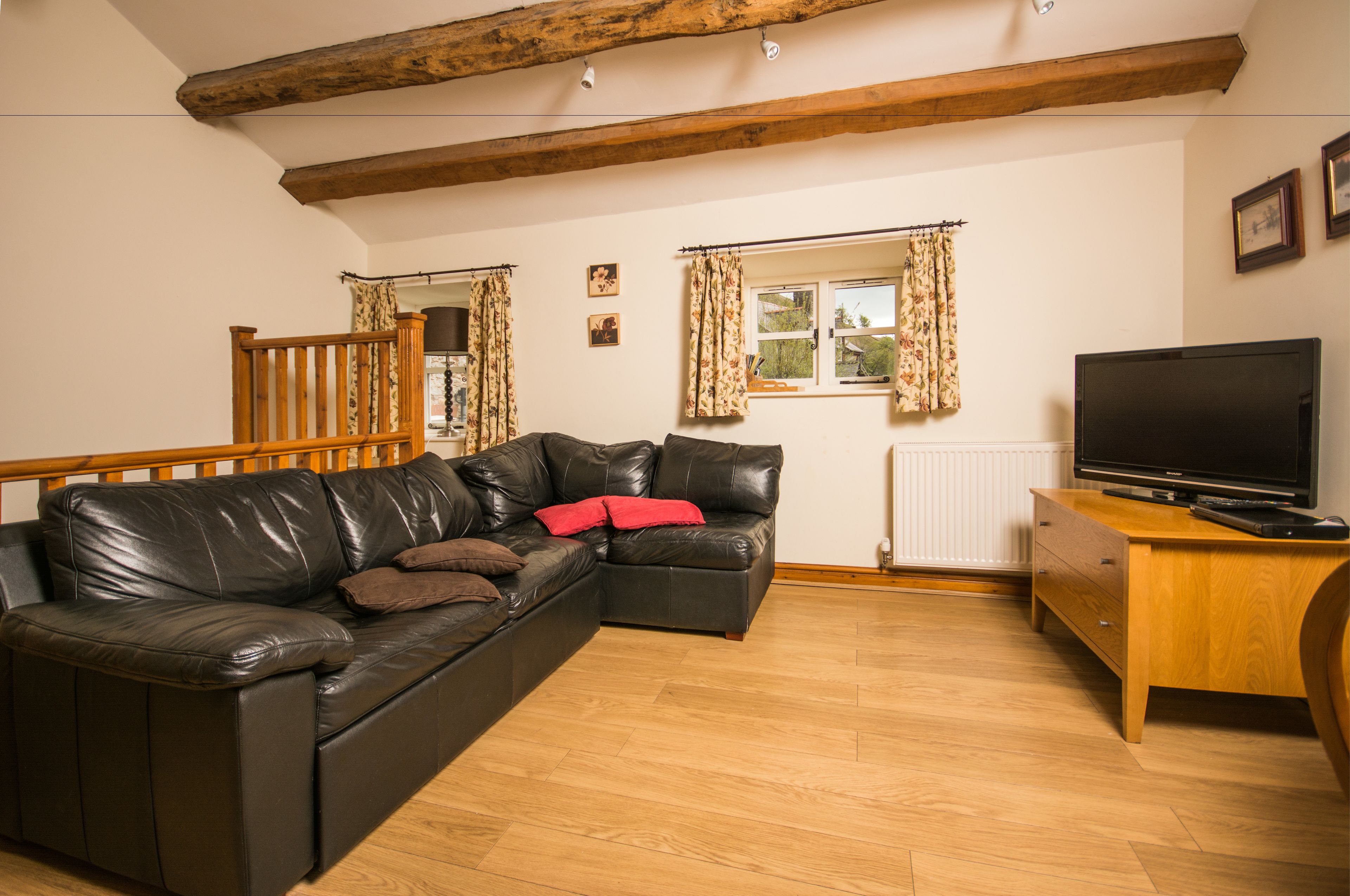 A living room with a black leather sofa, wooden flooring, a wooden TV stand, and a window with curtains.