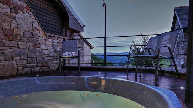 A hot tub with a view of the mountains, stone walls, and two chairs on a deck. - Panorama Cottages (Llangollen)