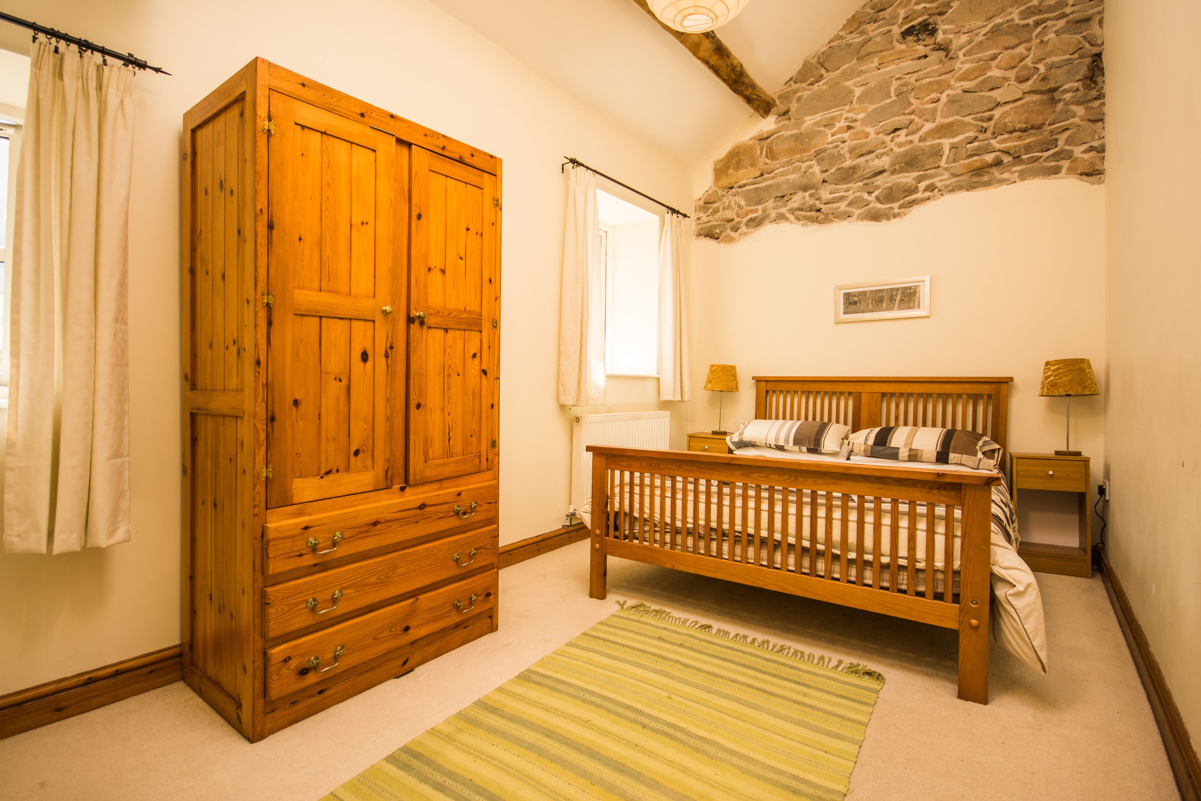 A bedroom with a wooden wardrobe, a bed with a headboard, a stone wall, a window with curtains, and a bedside table with a lamp.