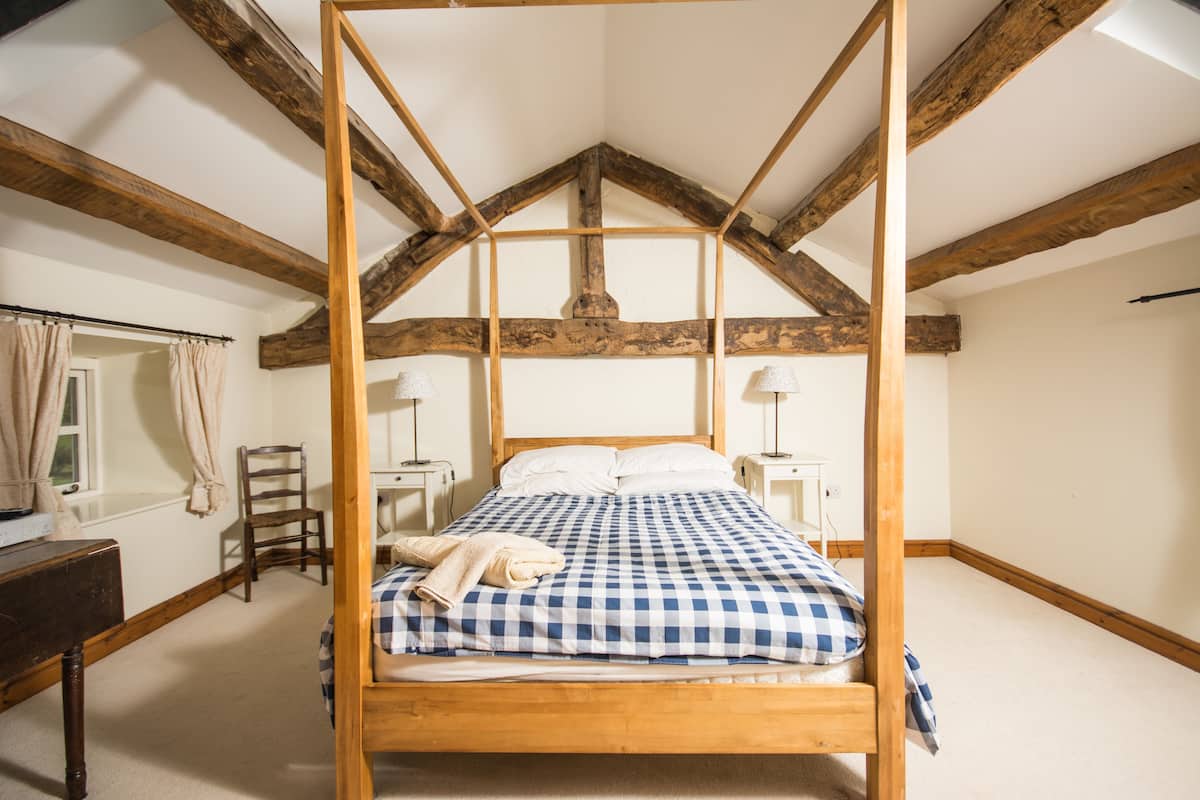 A four-poster bed with a checkered bedspread, wooden beams, and a window with curtains.
