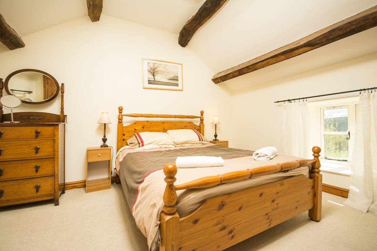 A bedroom with a wooden bed, a dresser, a nightstand, a mirror, and a window with curtains.