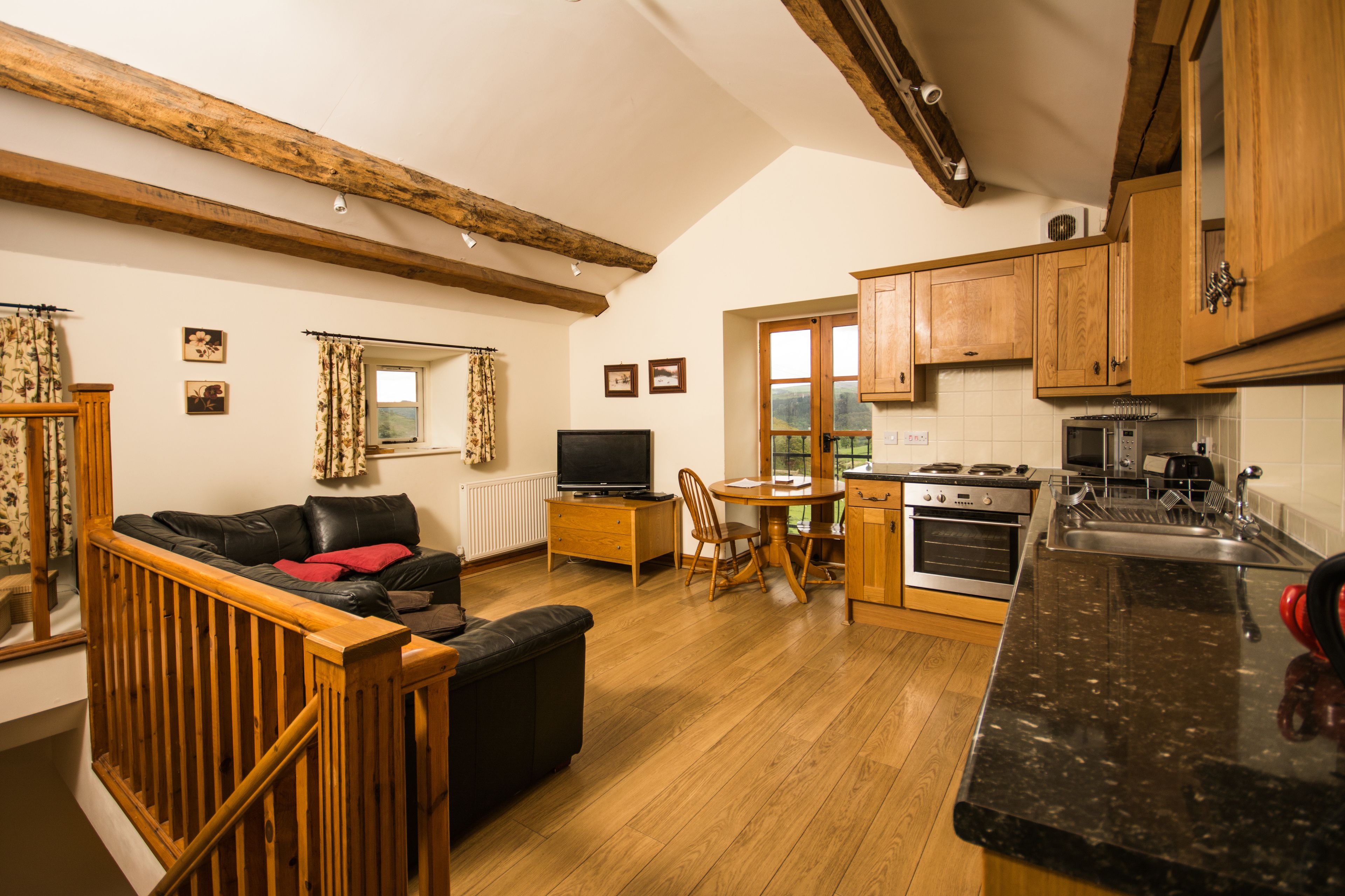 A cozy living room with a sofa, TV, and dining area. It features wooden beams, a kitchen with a stove, and a large window.