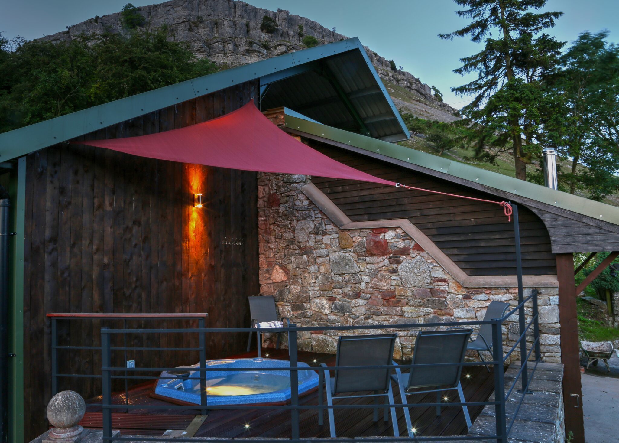 Una cabaña rústica con pared de piedra, terraza de madera, jacuzzi y dos sillas.