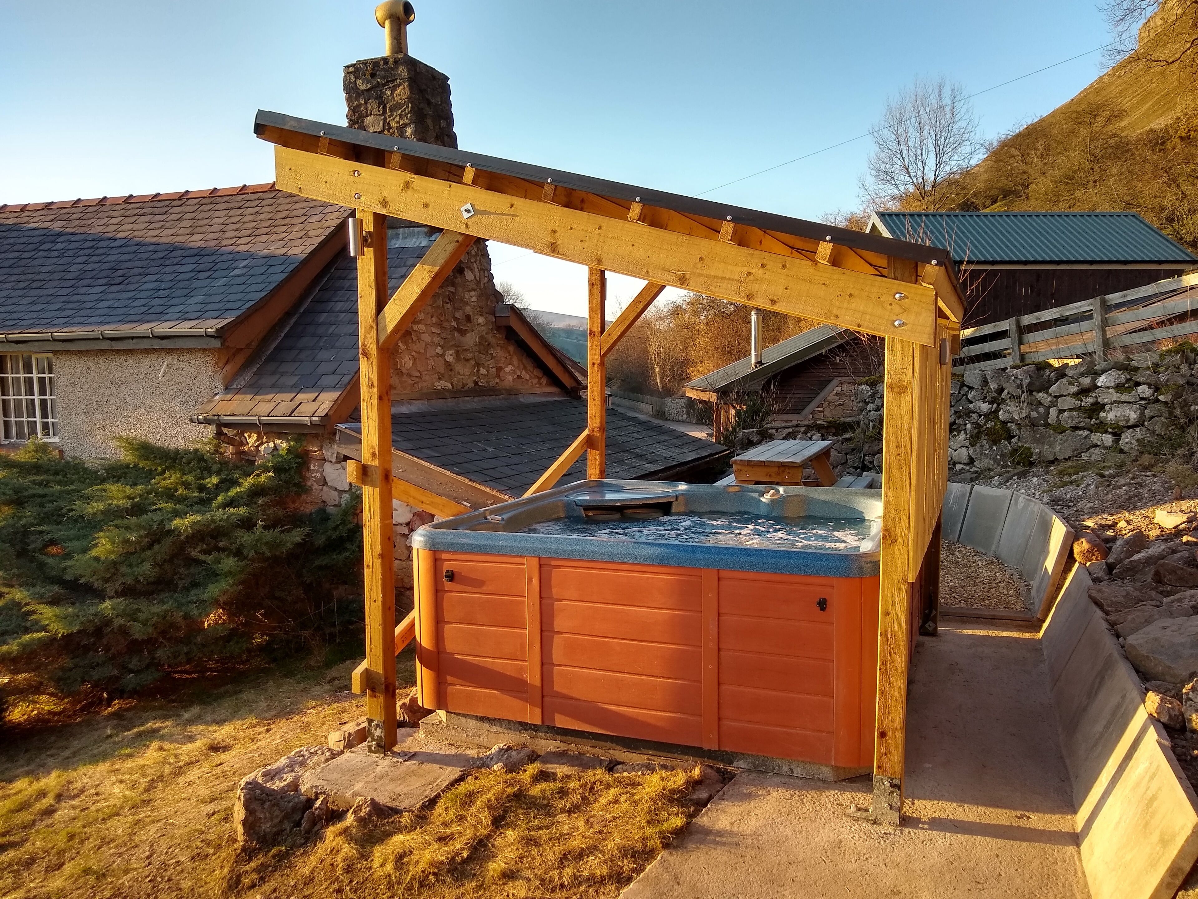Et spabad under et træbeklædt pergola, placeret i en have med en stenvæg og et træhegn i baggrunden.