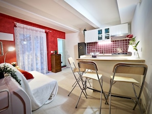 Un espacio habitable compacto con kitchenette, un comedor con sillas plegables, una cama con cubre cama blanco y una pared de acento roja.