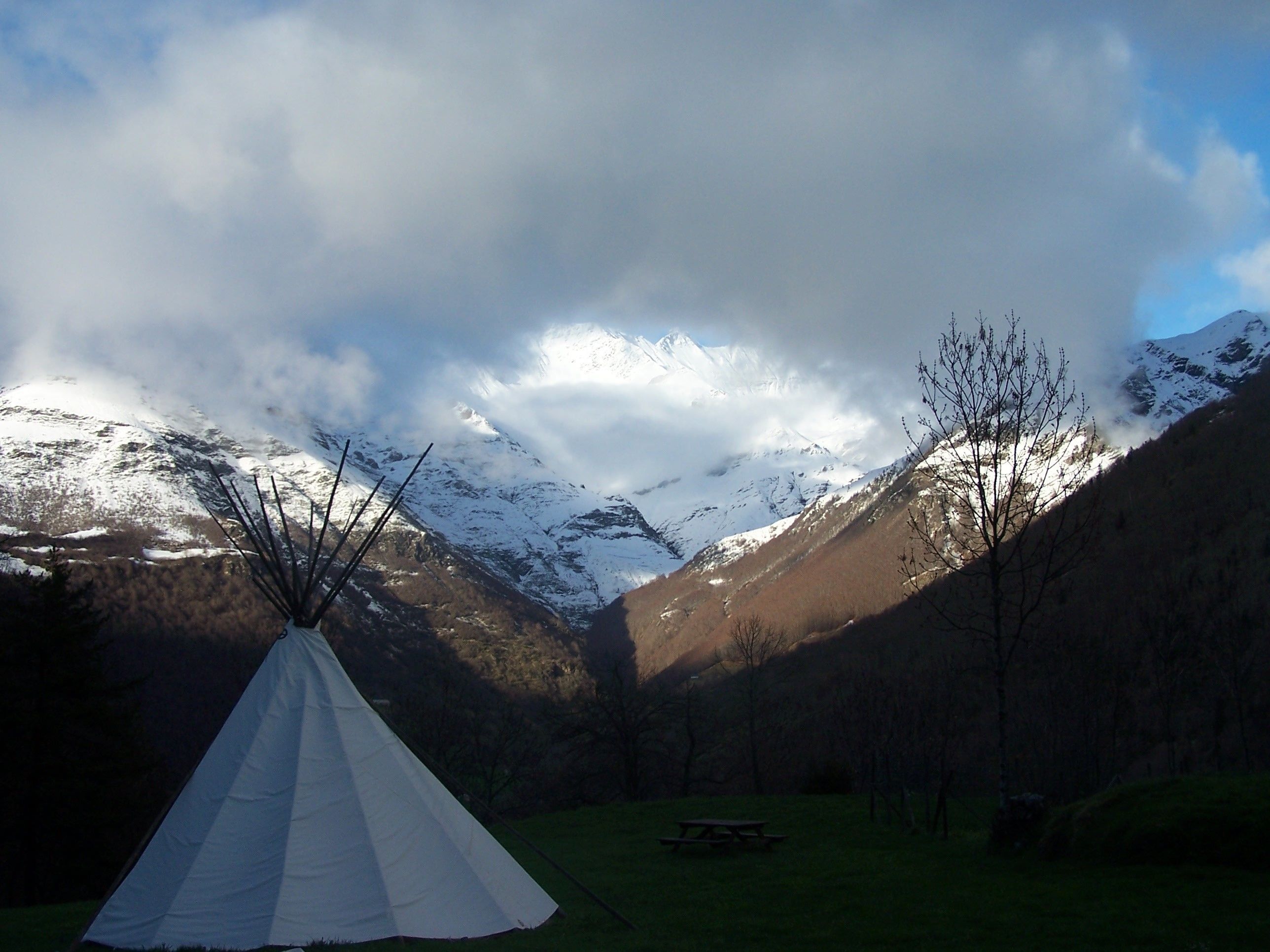 A white teepee in a valley with a picnic table, surrounded by mountains and trees.