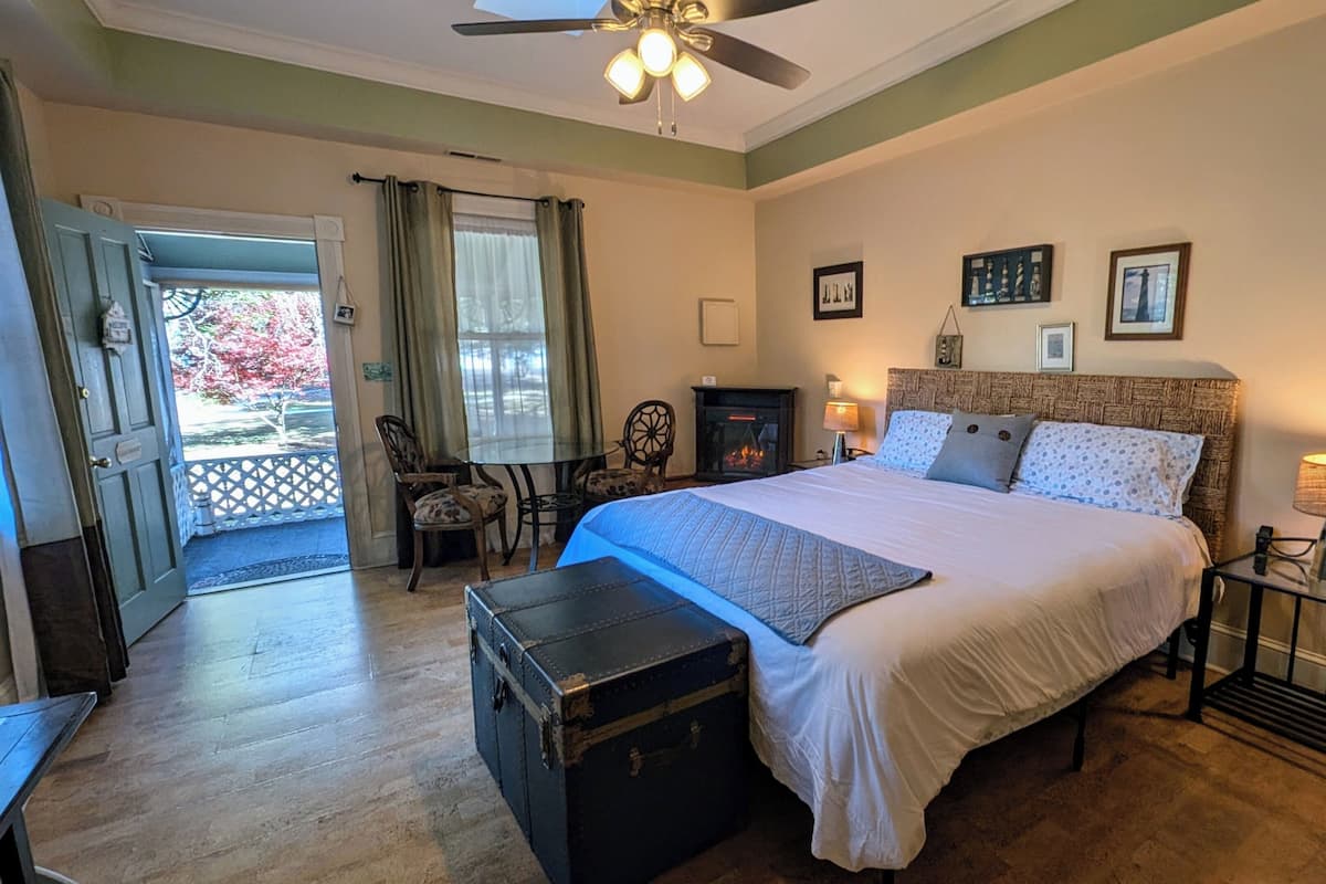 A bedroom with a bed, a fireplace, a ceiling fan, a nightstand with a lamp, and a suitcase.