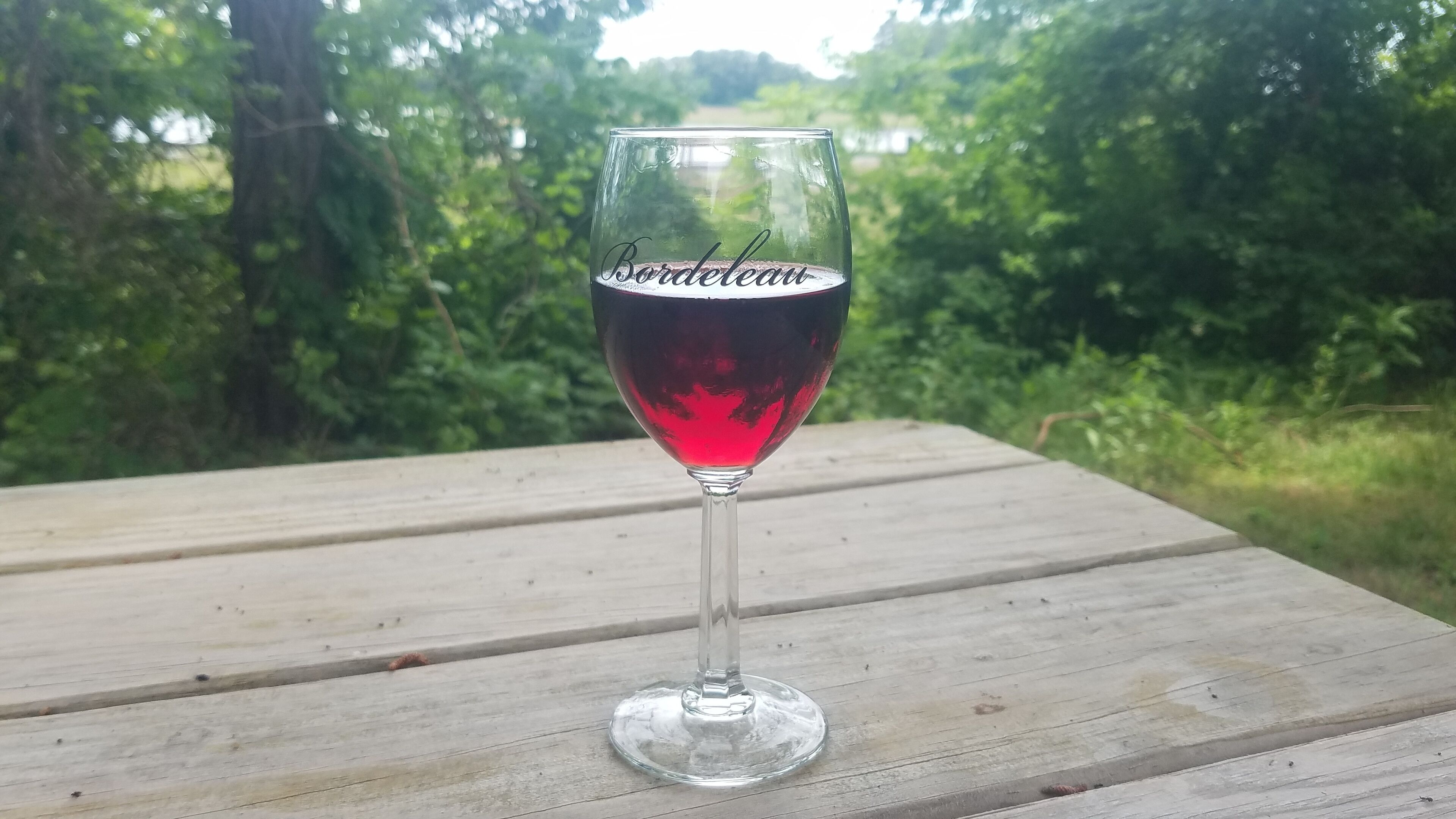 Een glas rode wijn met de aanduiding 'Bordeaux' op een houten tafel buiten.