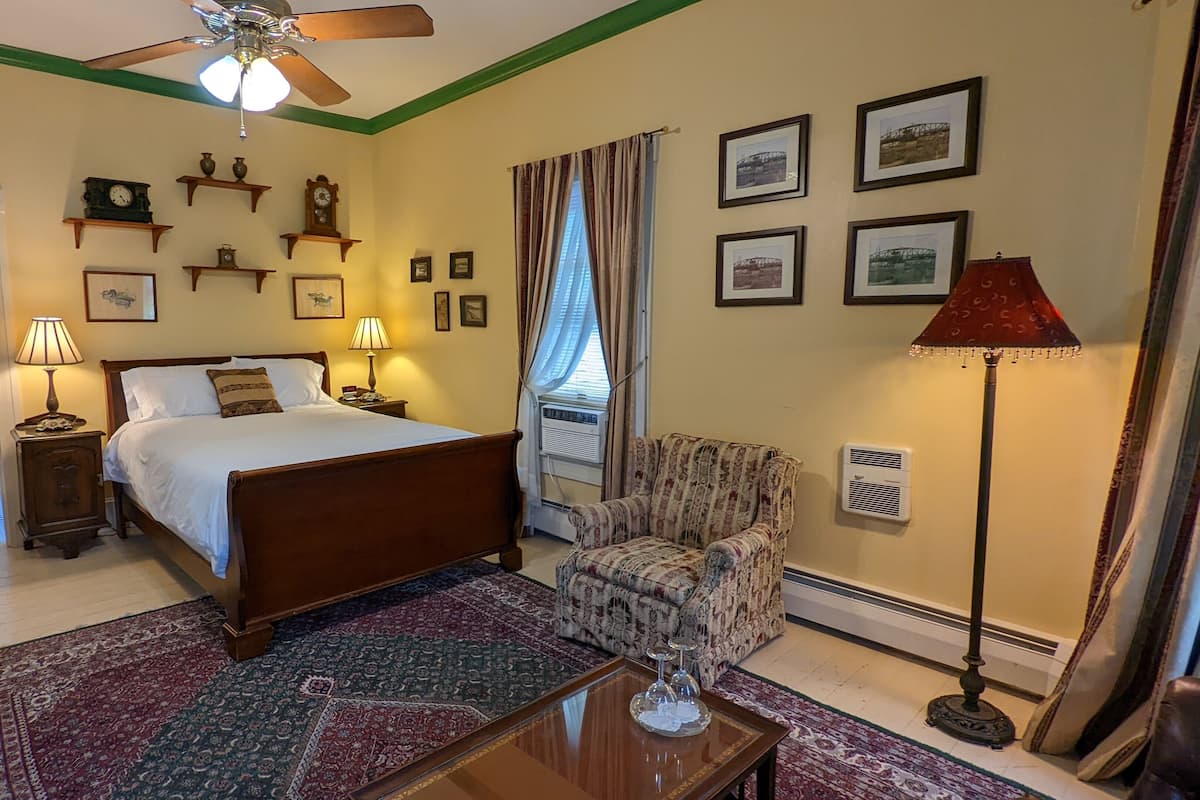 A bedroom with a bed, a chair, a table, a lamp, a ceiling fan, a window with curtains, and wall-mounted clocks and pictures.