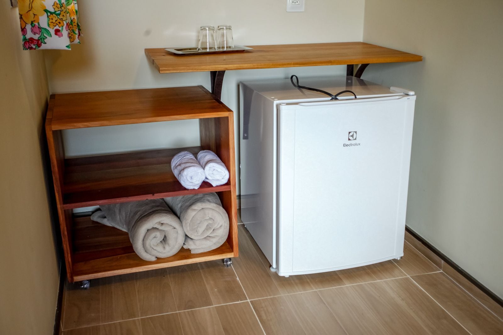 A compact room with a white refrigerator, a wooden shelf with towels, and a small table with glasses and a tray.
