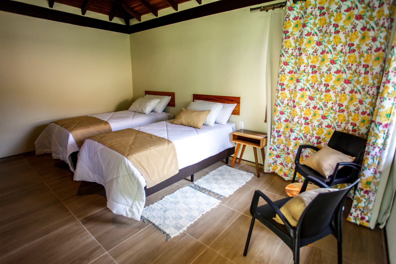 A room with two beds, a chair, a small table, and a window with floral curtains.