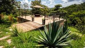 A patio with a wooden deck, a seating area with chairs and a table, surrounded by lush greenery and a view of a hilly landscape.