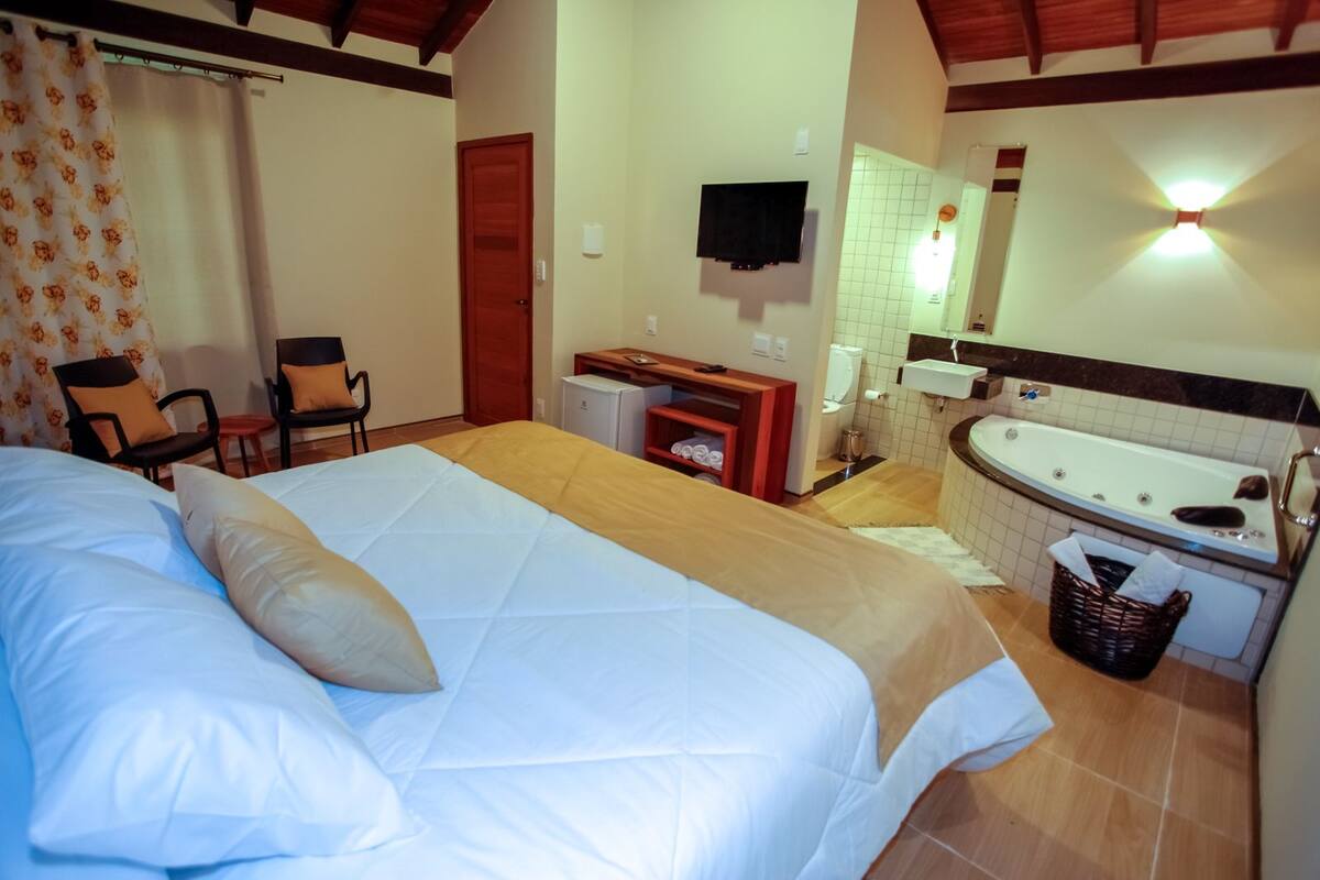 A hotel room with a bed, chairs, a desk, a TV, a bathroom with a bathtub, and a tiled floor.