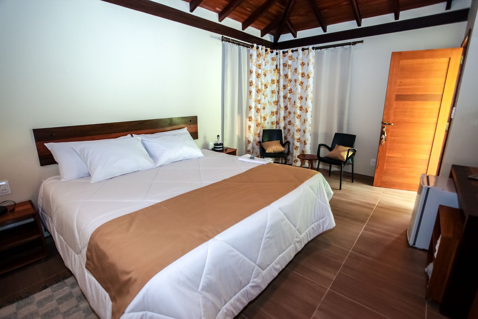 A hotel room with a large bed, two chairs, a wooden door, and a tiled floor.