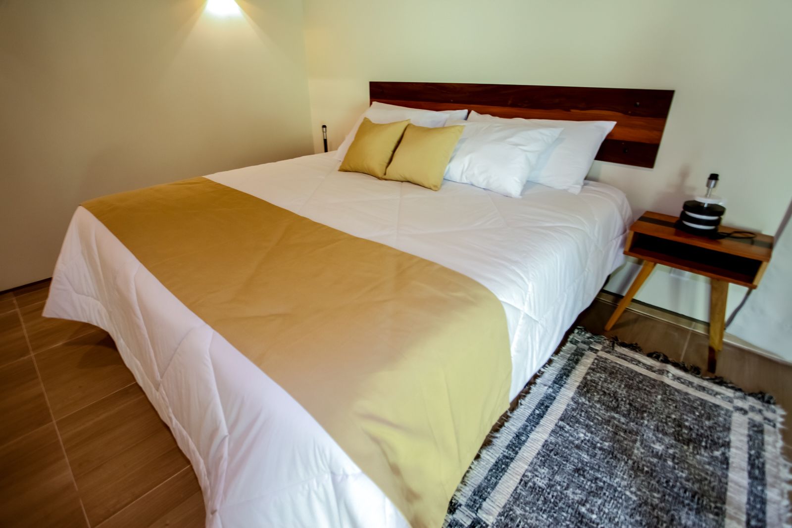 Una cama bien tendida con cubrecama blanco y beige, un cabecero de madera y una mesita de noche pequeña con una lámpara y un objeto negro.