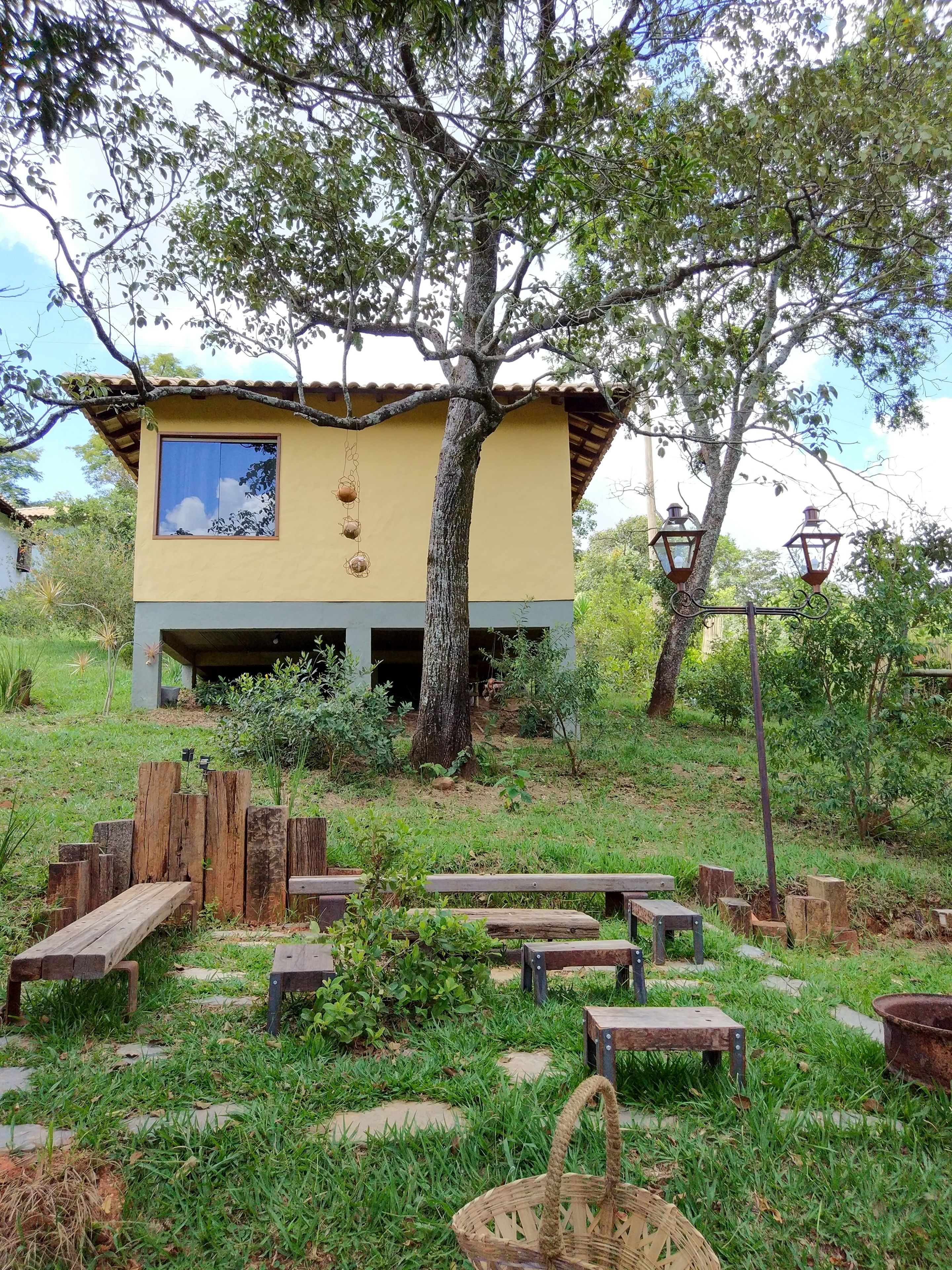 Un chalet rustique doté d’une grande fenêtre, entouré d’un jardin avec des bancs en bois et un arbre.