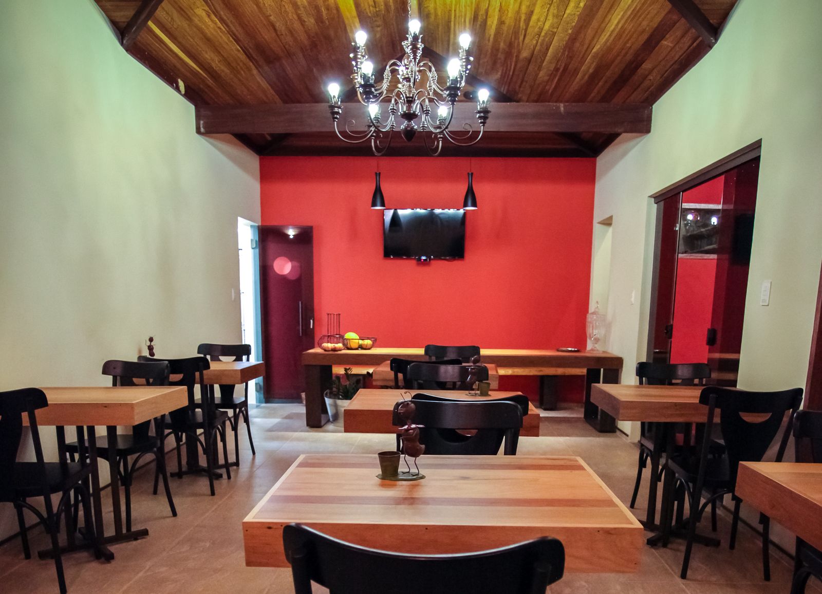 A dining area with wooden tables and chairs, a chandelier, and a flat-screen TV mounted on the red wall.
