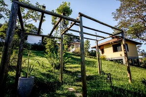 Uma construção rústica de madeira num jardim exuberante, com uma casa amarela ao fundo.