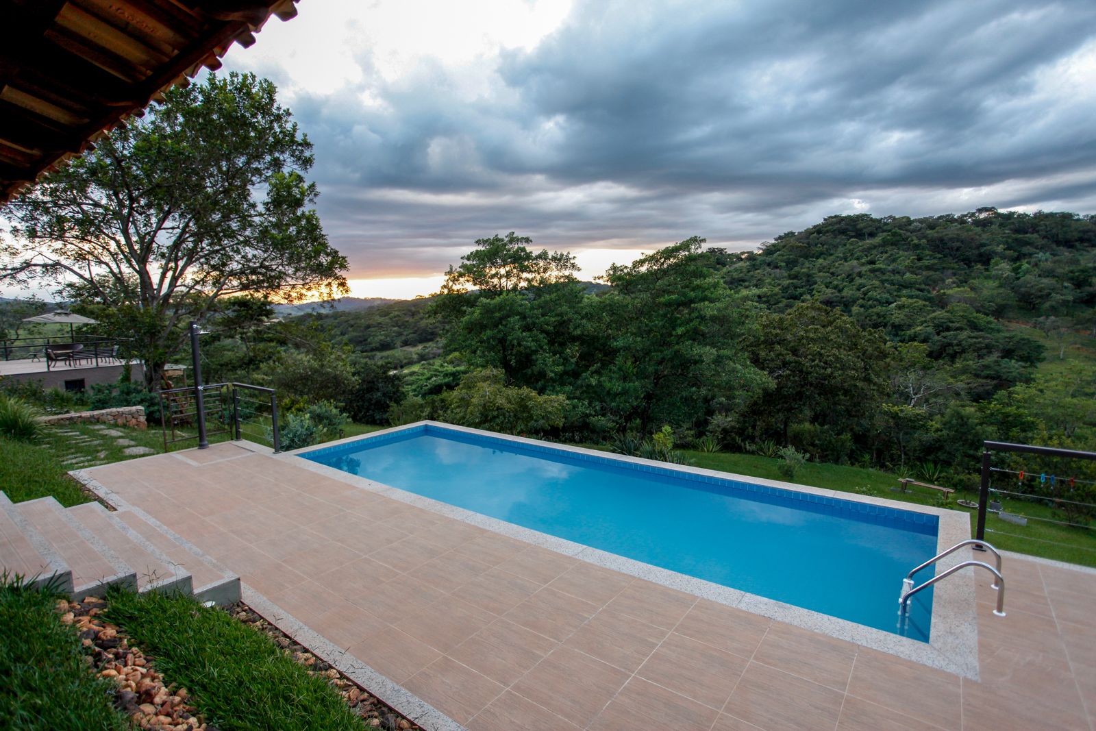 Una piscina rectangular con solárium de baldosas, rodeada de vegetación exuberante y árboles, bajo un cielo nublado.