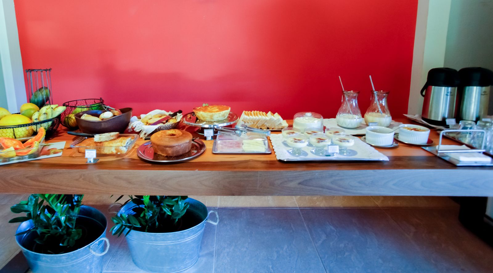 A breakfast buffet with various food items and beverages on a wooden table against a red wall.