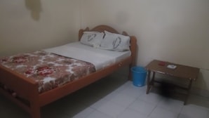 Una cama individual con colcha estampada, cabecera de madera y dos almohadas. Junto a la cama, una mesita de noche de madera con un cubo de basura azul.