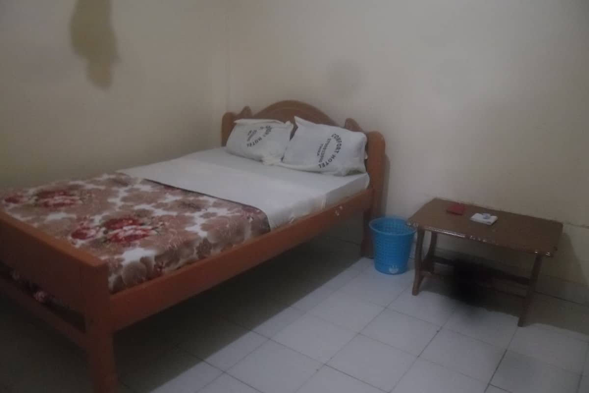 Una cama individual con colcha estampada, cabecera de madera y dos almohadas. Junto a la cama, una mesita de noche de madera con un cubo de basura azul.