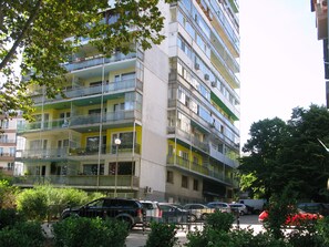 Um edifício residencial com vários andares, varandas, área de estacionamento e vegetação ao redor.