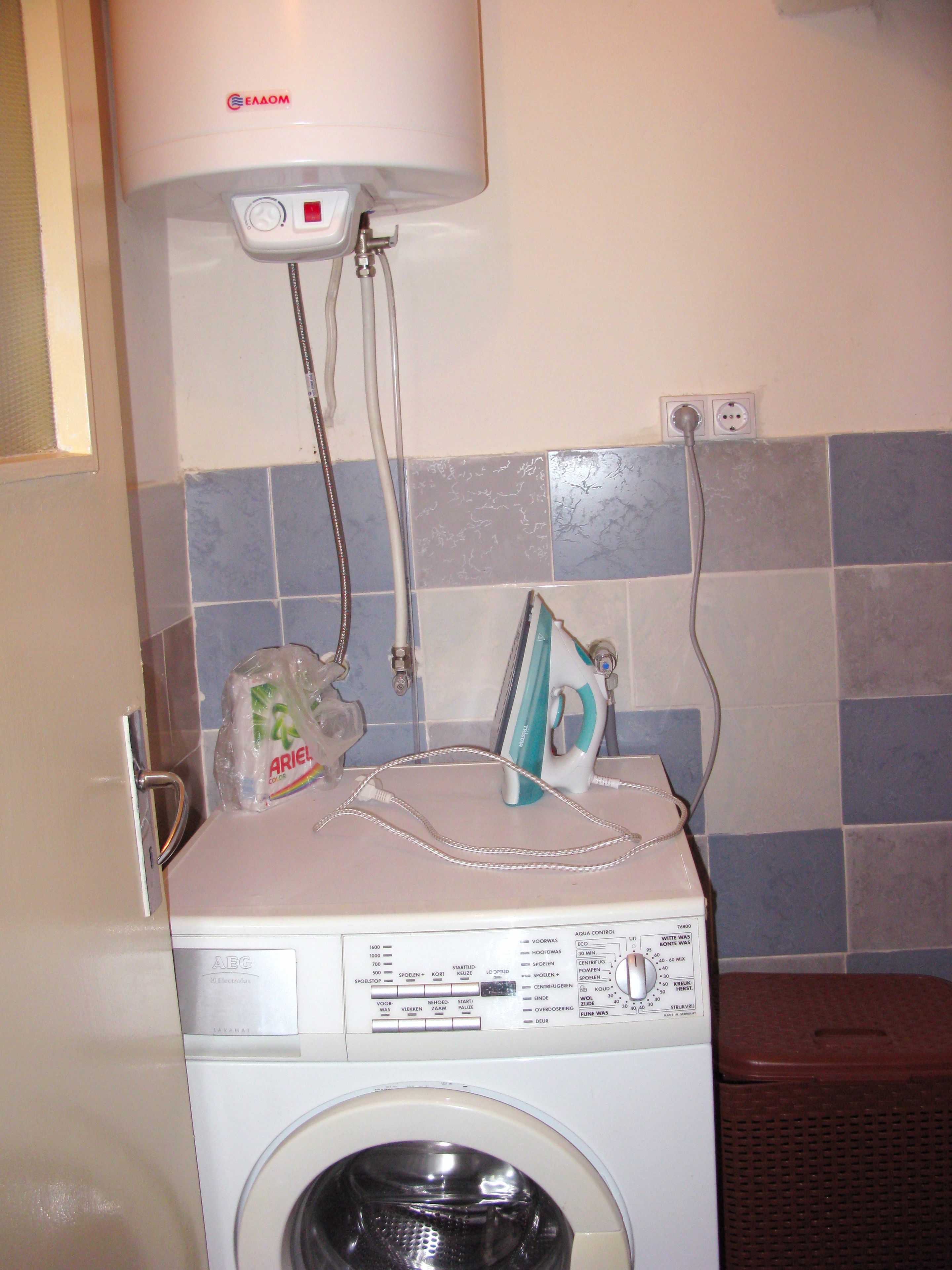 A front-loading washing machine with a control panel, a water heater, and a power outlet on the wall.