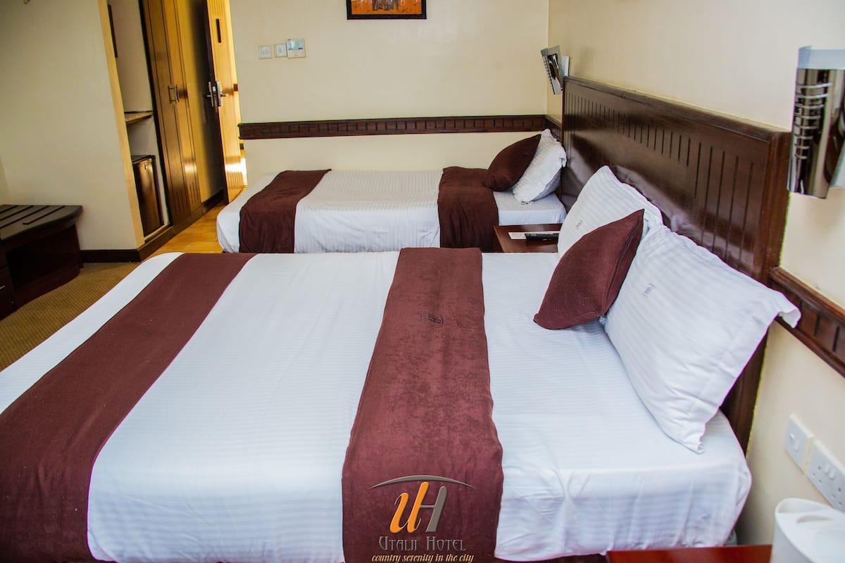 A hotel room with two beds, each with white linens and brown headboards. There is a wooden wardrobe and a nightstand with a lamp.
