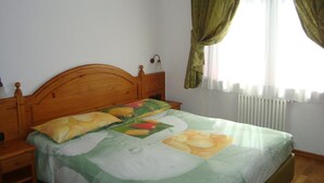 A bedroom with a wooden headboard, a bed with a green and white floral bedspread, a nightstand, a wall-mounted lamp, and a window with green curtains. - La Falconetta (Ayas)