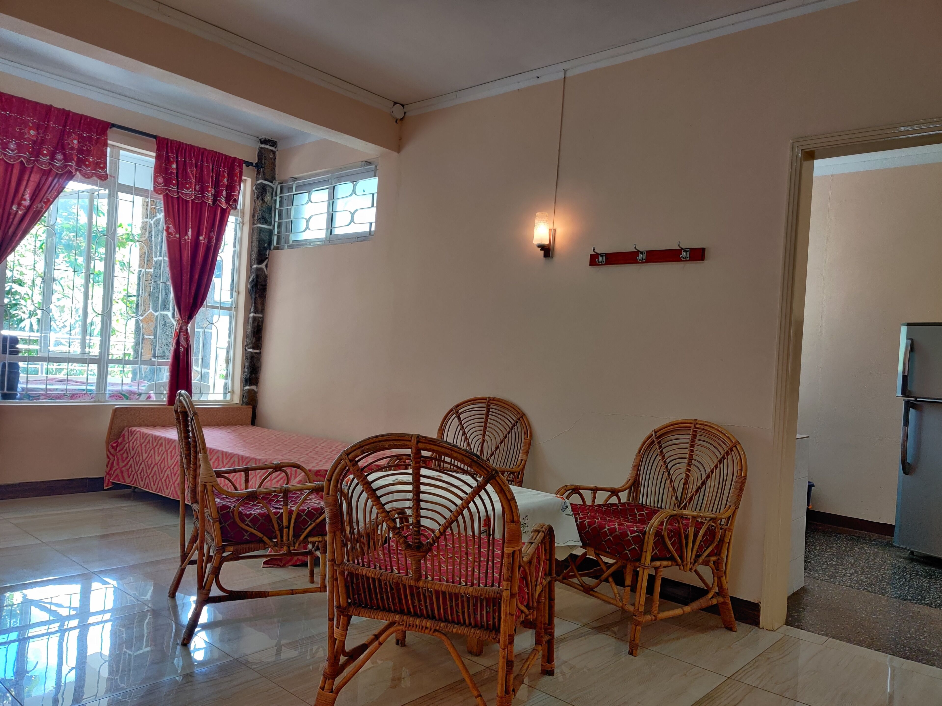 A dining area with wicker chairs and a table, a window with curtains, a wall-mounted light, and a refrigerator in the corner.