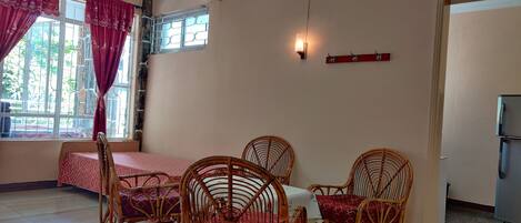 A dining area with wicker chairs and a table, a window with curtains, a wall-mounted light, and a refrigerator in the corner.