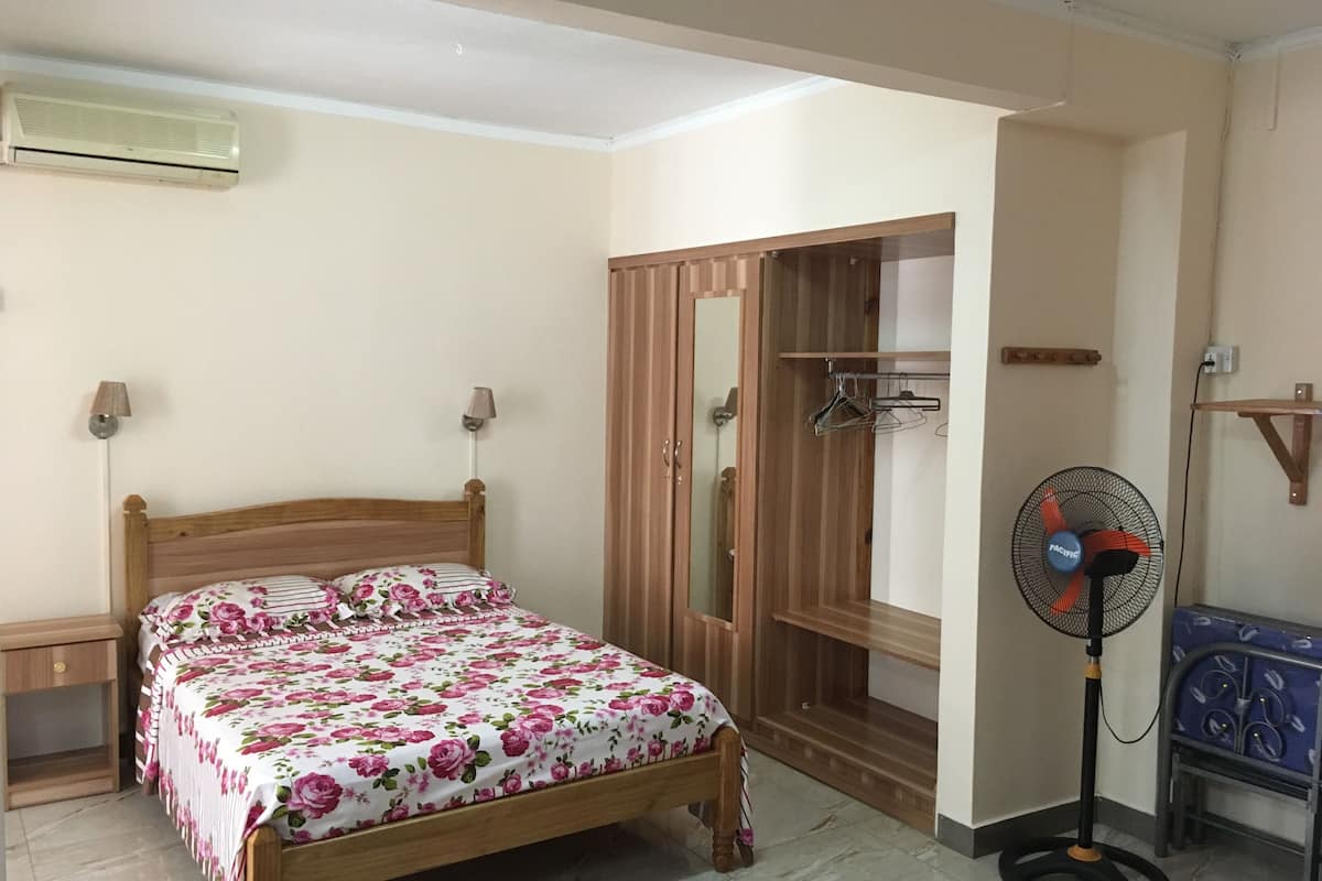 Une chambre avec un lit en bois, une armoire, un ventilateur sur pied et une petite table.