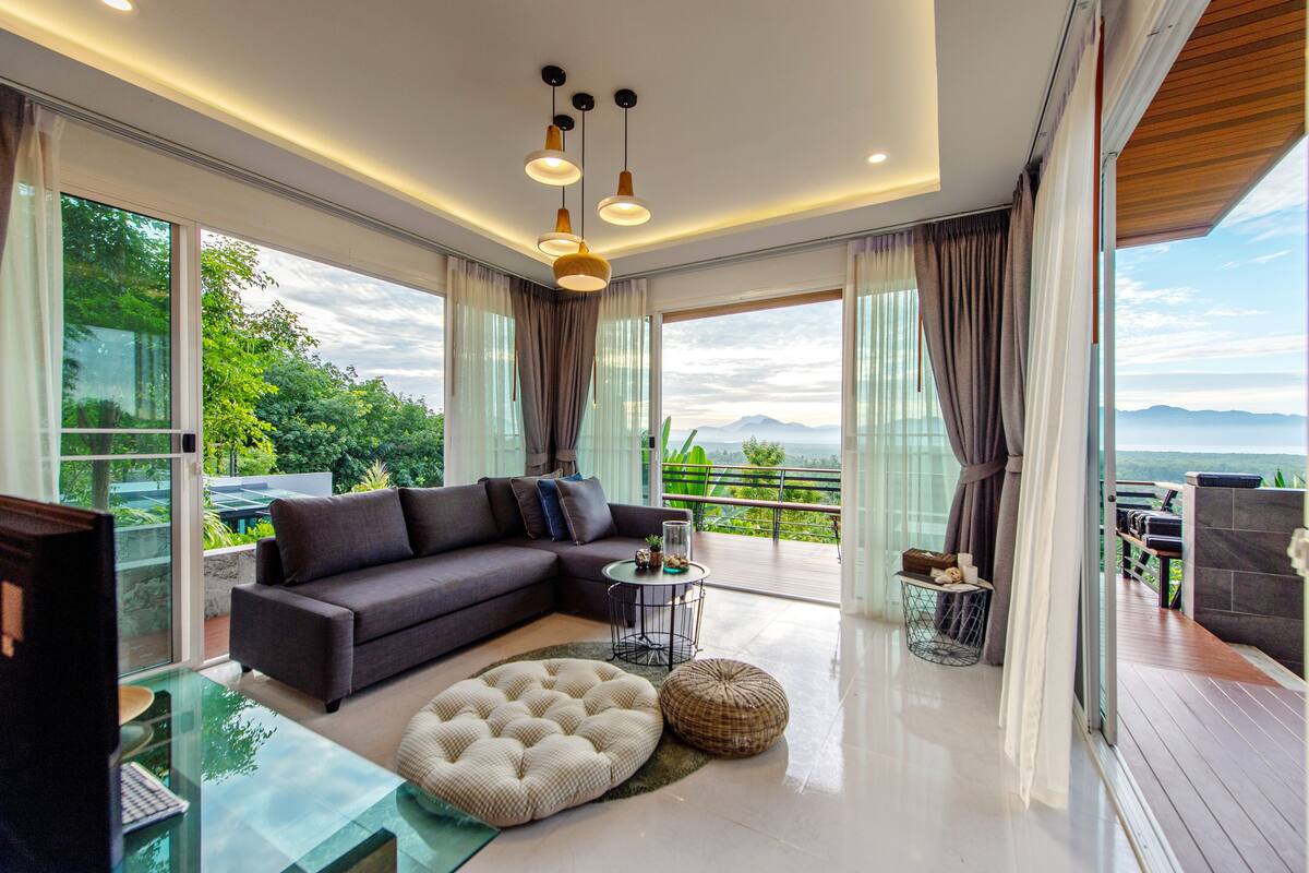 Uma sala de estar moderna com um sofá seccional, uma mesa de centro de vidro e um banquinho redondo. Há uma varanda com vista para vegetação e montanhas.