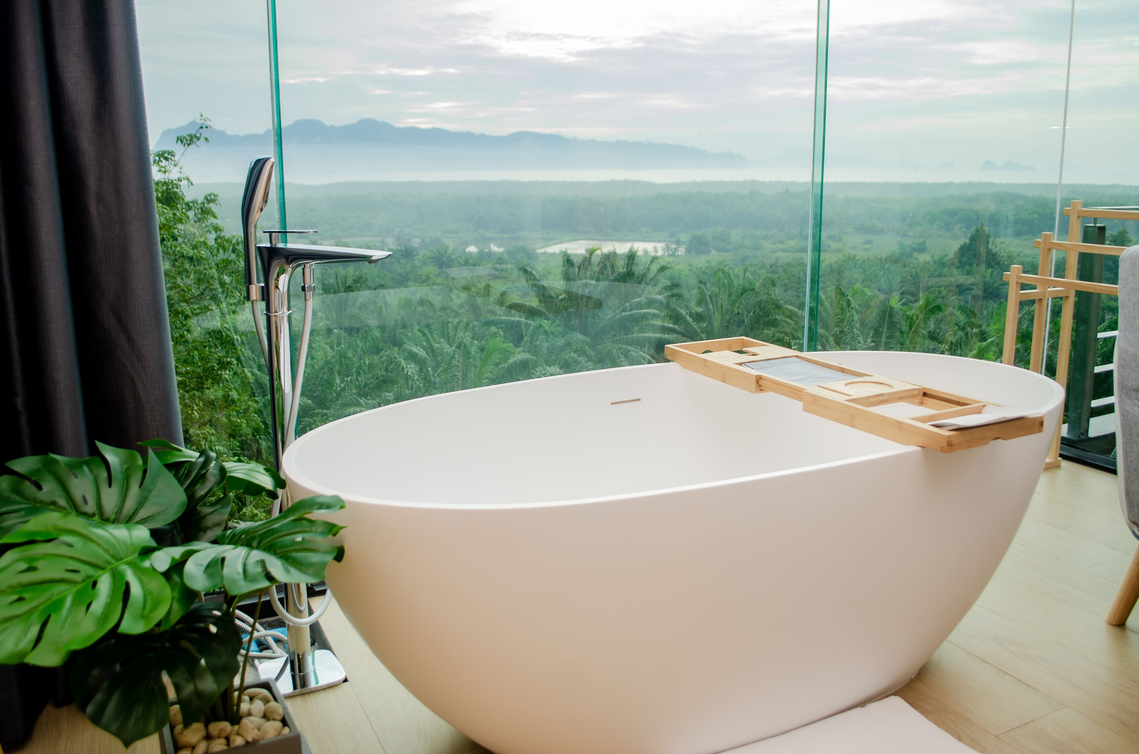 A modern bathroom with a freestanding bathtub, a large window offering a view of a lush landscape, and a potted plant.