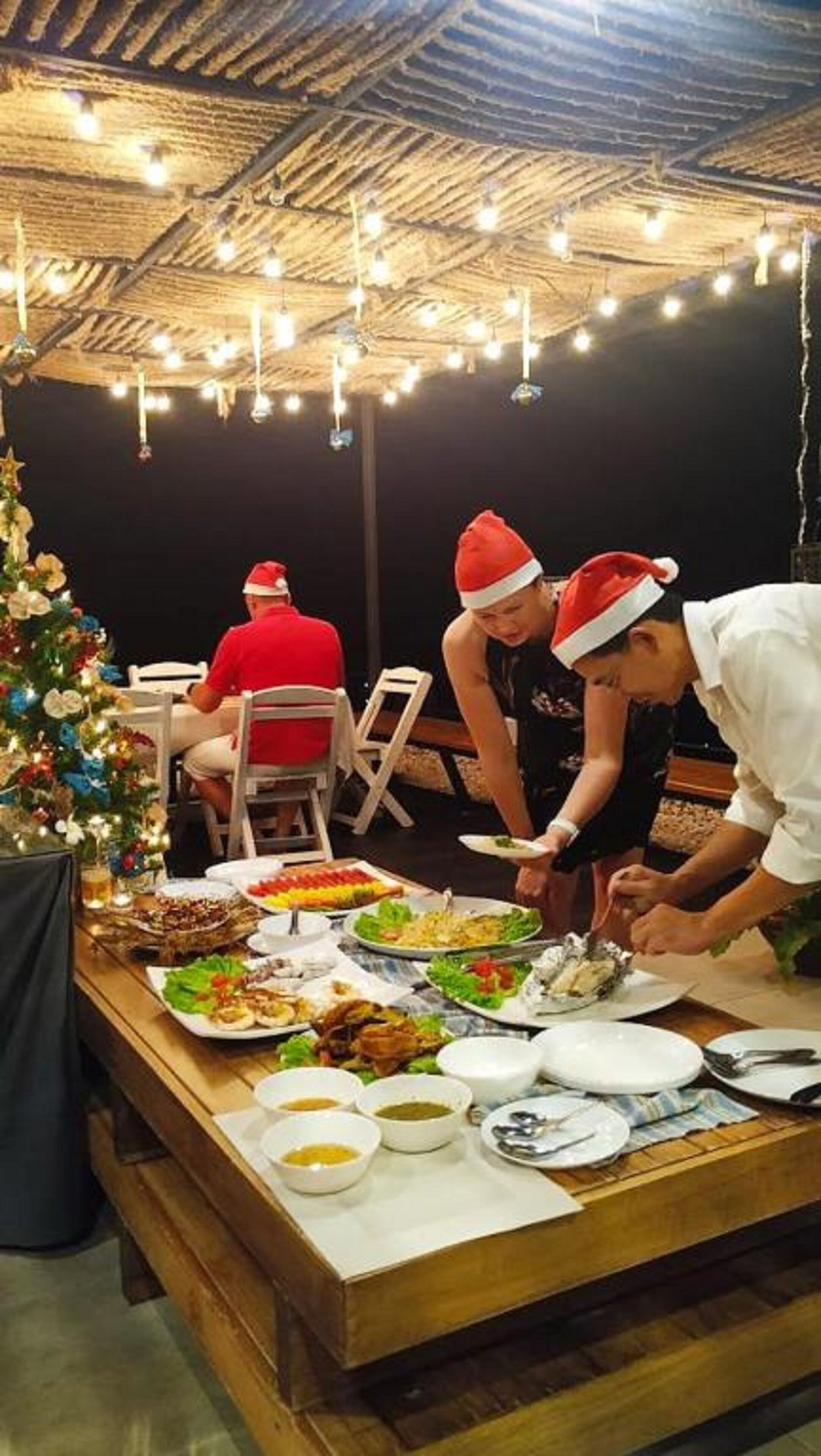 A dining area with a wooden table set for a meal, a Christmas tree, and string lights on the ceiling.