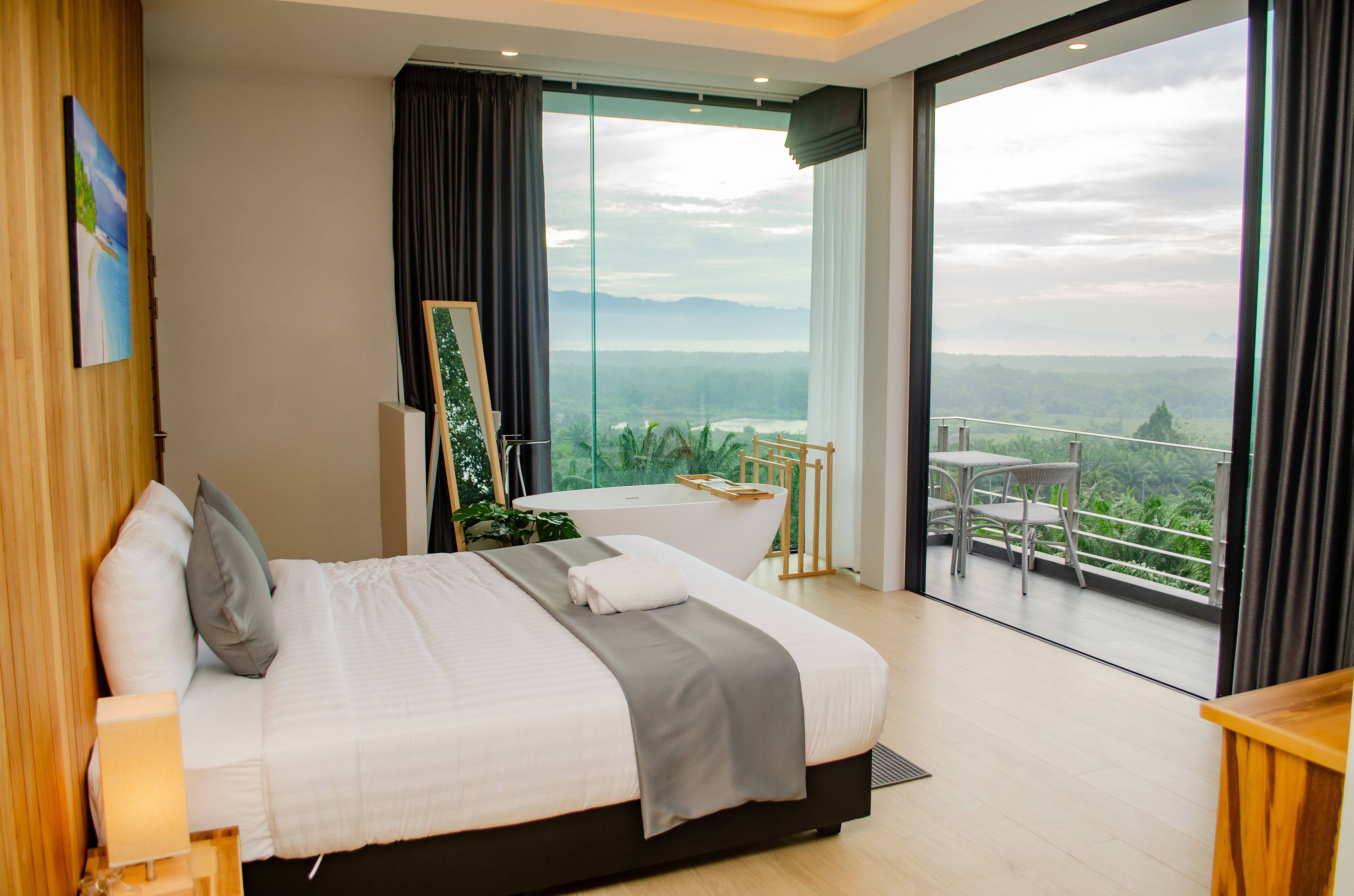A modern hotel room with a large bed, a painting on the wall, a view of a balcony with outdoor furniture, and a large window.