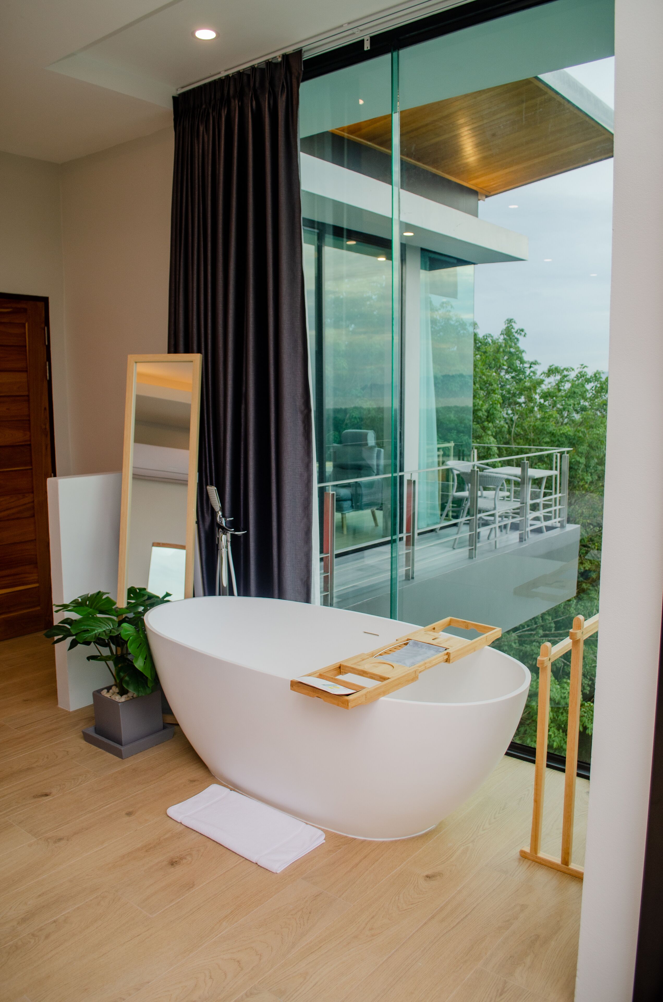 A modern bathroom with a freestanding bathtub, a large mirror, and a view of a balcony with outdoor furniture.