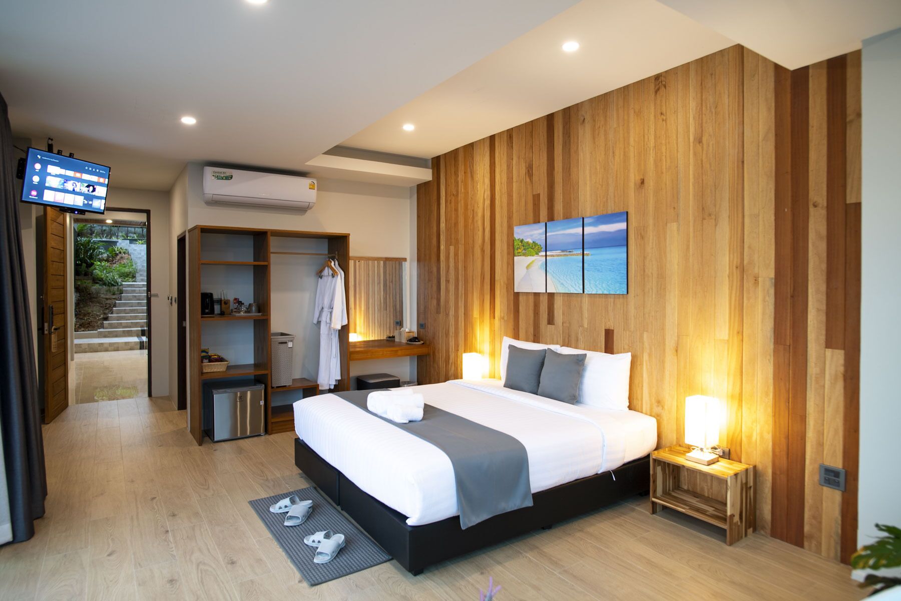 A modern hotel room with a wooden accent wall, a large bed, a bedside table with a lamp, a wardrobe, and a TV in the corner.