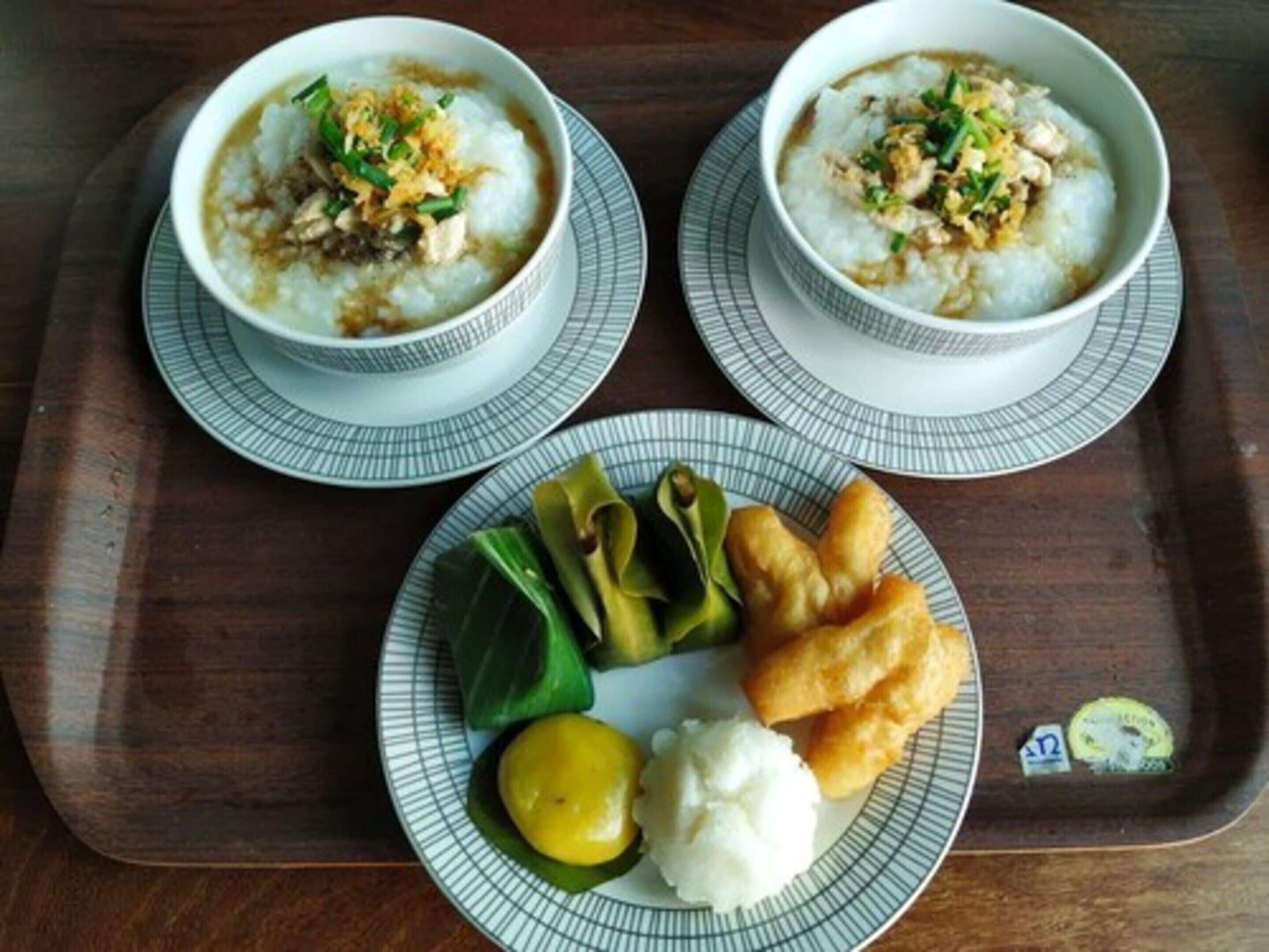 Two bowls of rice porridge with toppings, a plate with vegetables and fried items, and a small dish of white sauce.