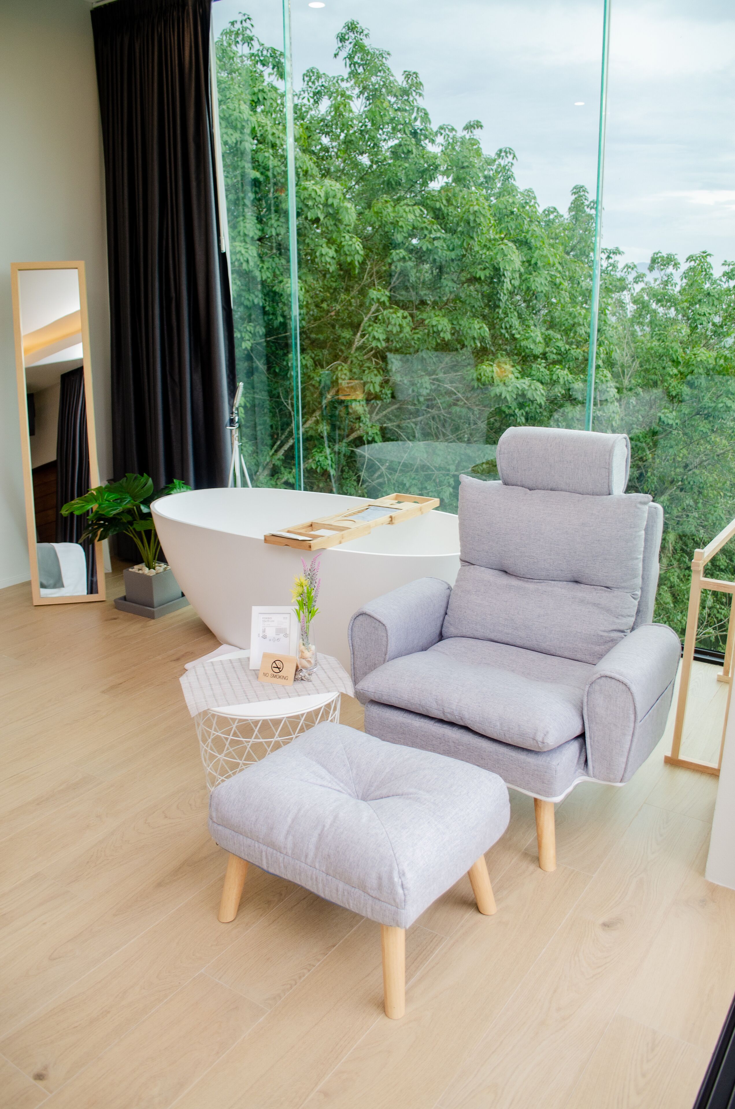 Una habitación moderna con un ventanal grande, una bañera, un sillón gris y una pequeña mesa redonda con una planta.