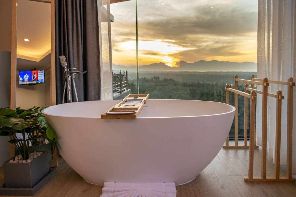Um banheiro moderno com uma banheira grande, uma mesinha de madeira, uma planta em vaso e vista para um pôr do sol sobre montanhas.