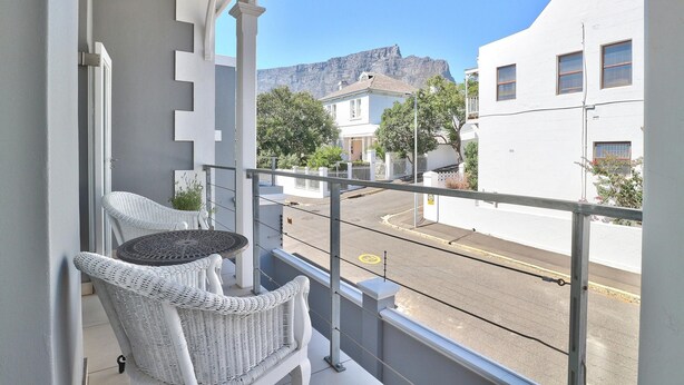 A balcony with wicker chairs and a small table, overlooking a street with buildings and a mountain in the background. - Maudville (Cape Town)
